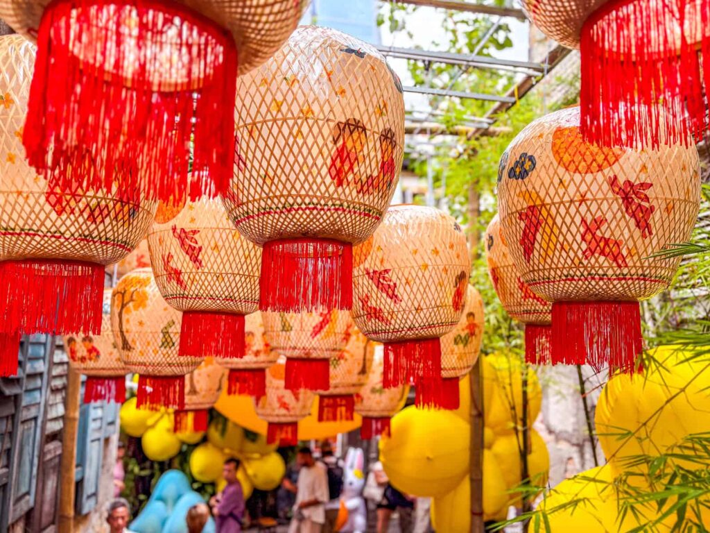 Wiszące tradycyjne chińskie lampiony z czerwonymi frędzlami i rysunkami królików nad alejką Kwai Chai Hong. W tle widać rozmyte postacie turystów i żółte dekoracje.