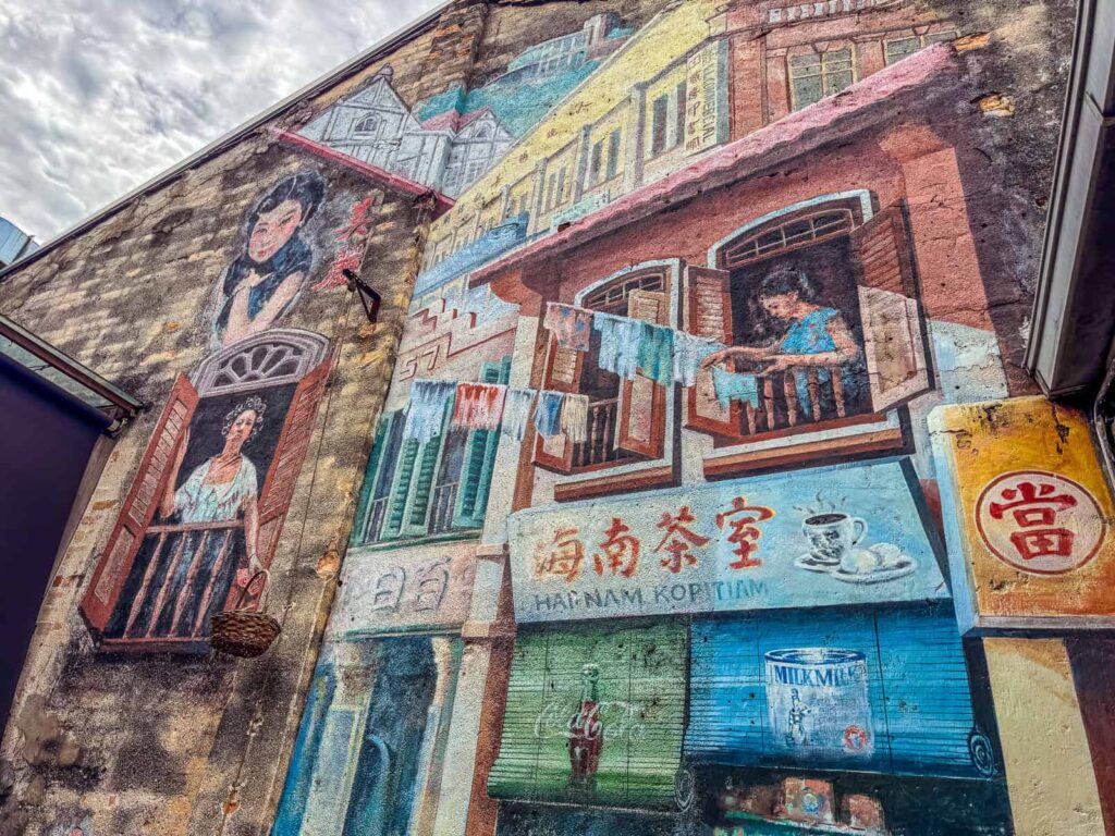 Wielkoformatowy mural na ścianie budynku w Kwai Chai Hong. Przedstawia przekrój życia w dawnej dzielnicy: dzieci skaczące przez skakankę, klientów w tradycyjnej kawiarni Hainam Kopitiam oraz kobietę wieszającą pranie i spuszczającą koszyk na sznurku z piętra.
