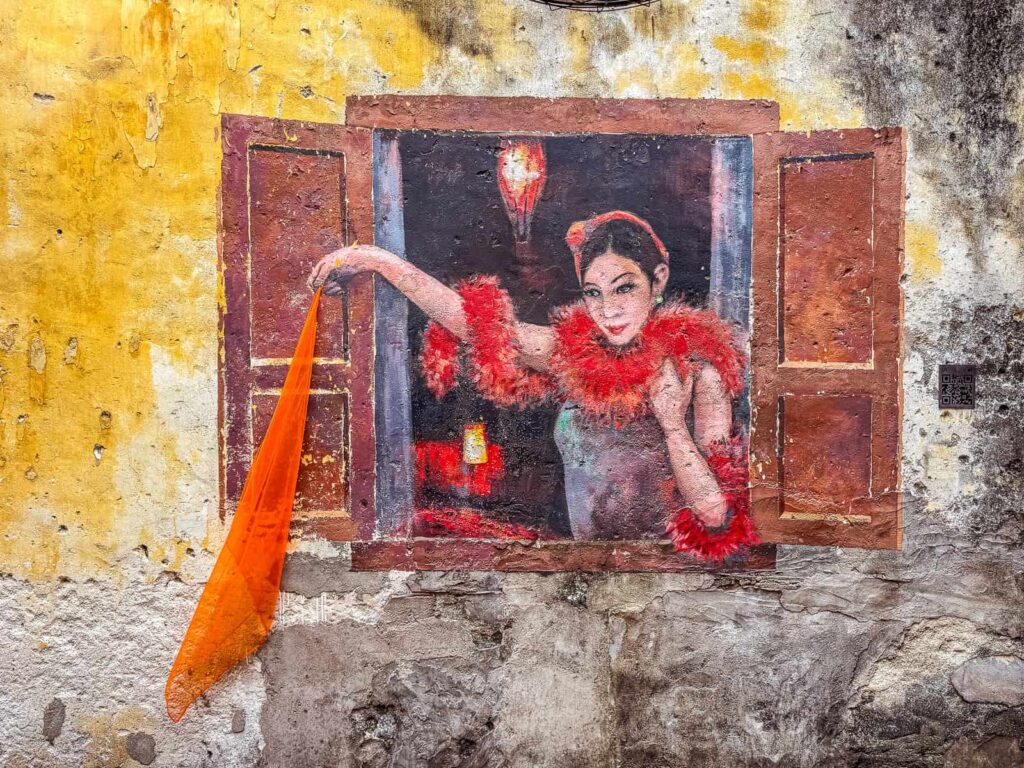 Mural na żółtej, zniszczonej ścianie w Kwai Chai Hong. Przedstawia kobietę w stylu lat 60. w czerwonym boa, wyglądającą przez namalowane okno. Kobieta trzyma w dłoni prawdziwy pomarańczowy szal, który zwisa ze ściany.