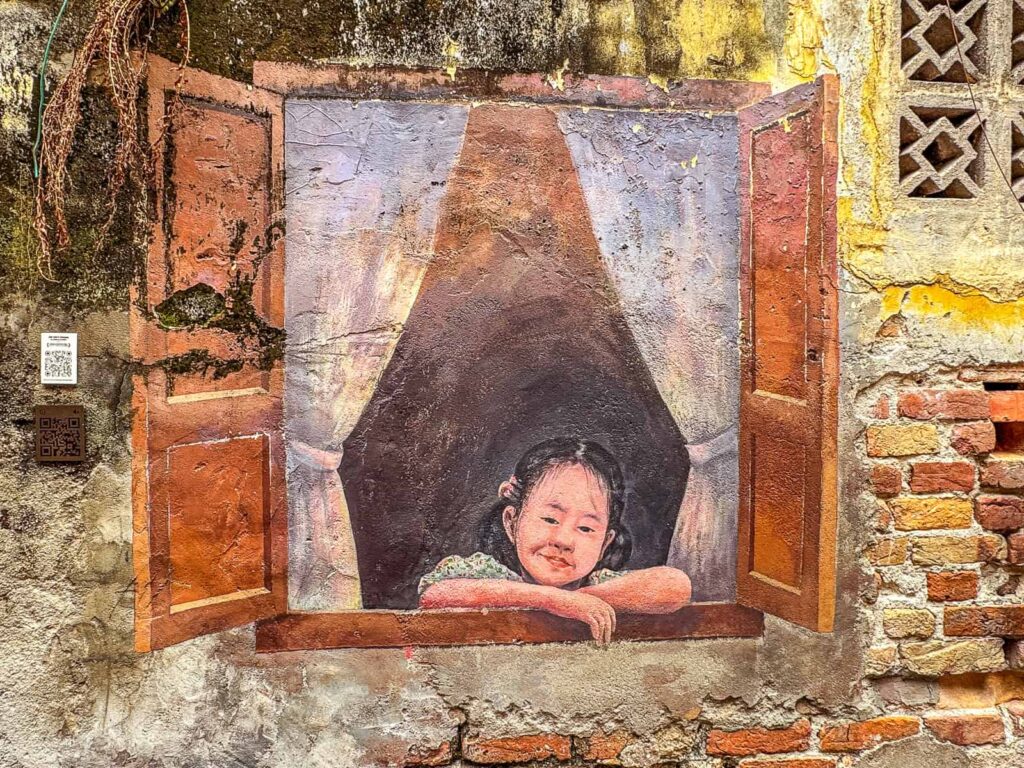 Mural na zniszczonej, częściowo ceglanej ścianie w Kwai Chai Hong, przedstawiający uśmiechniętą dziewczynkę wyglądającą przez namalowane okno z brązowymi okiennicami.