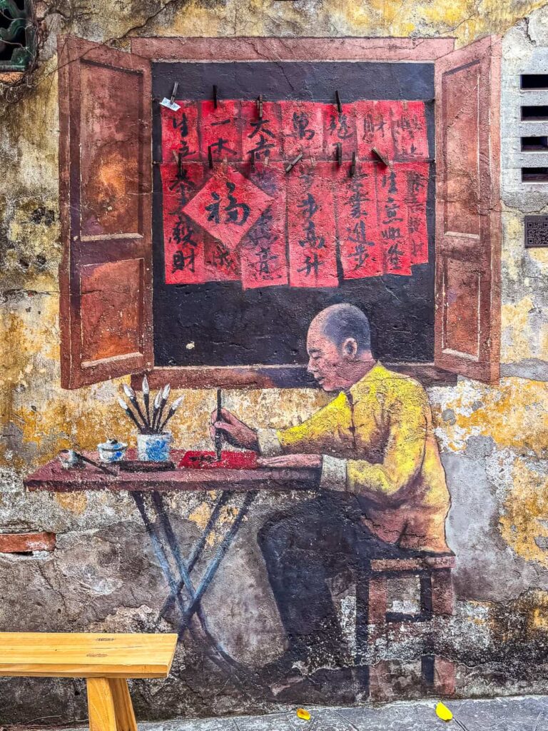 Mural w Kwai Chai Hong przedstawiający mężczyznę w żółtej koszuli, piszącego chińską kaligrafię przy małym stoliku. W tle namalowane jest otwarte okno z suszącymi się czerwonymi kartkami z pismem.