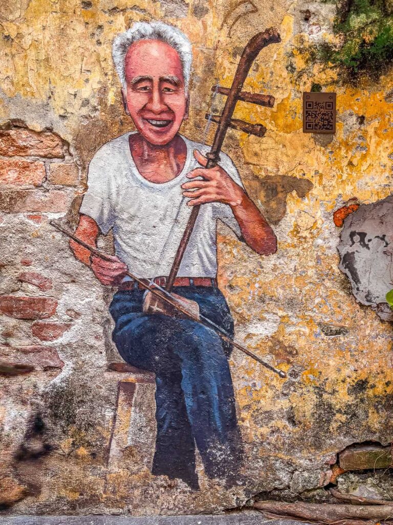 Mural na zniszczonej ścianie w Kwai Chai Hong przedstawiający uśmiechniętego starszego mężczyznę grającego na tradycyjnym chińskim instrumencie erhu. Obok widoczny jest kod QR do odsłuchania nagrania.