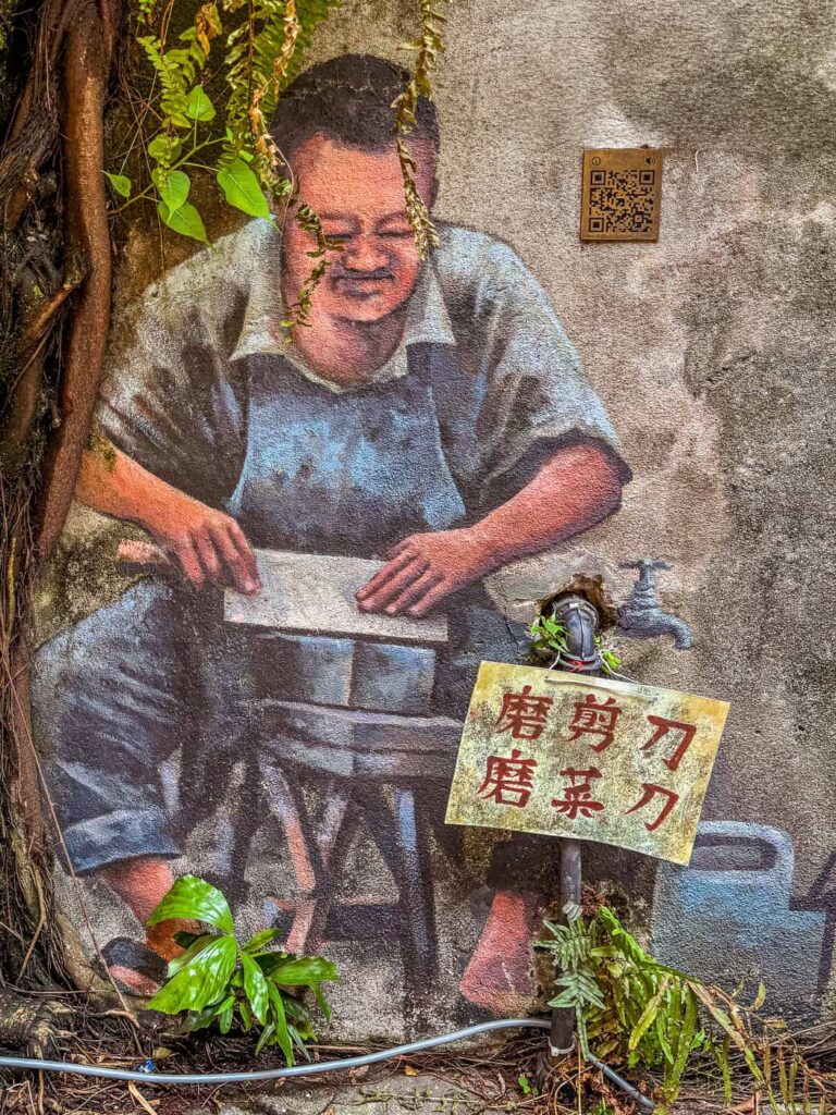 Mural w Kwai Chai Hong w Kuala Lumpur przedstawiający starszego mężczyznę ostrzącego noże. Obraz jest wkomponowany w ścianę, wykorzystując prawdziwą rurę z wodą i roślinność.